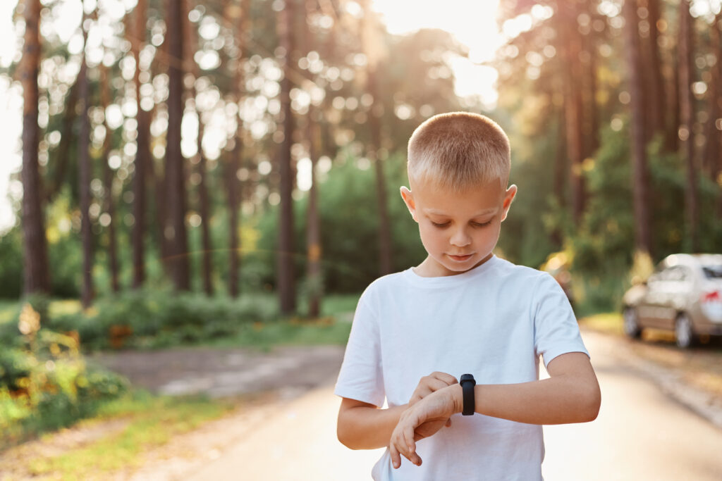 Little blonde boy using fitness bend touching button and touchscreen while posing outdoor in park, touching button and setting smart watch before running on road. Little blonde boy using fitness bend touching button and touchscreen while posing outdoor in park